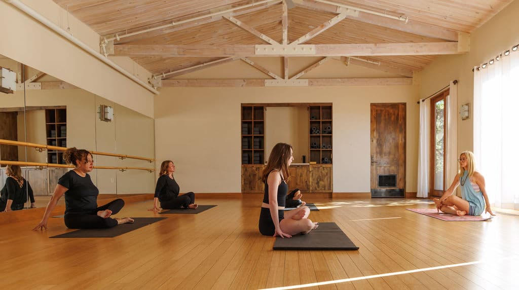 Four women sitting on yoga mats in a bright studio with high beamed ceilings.